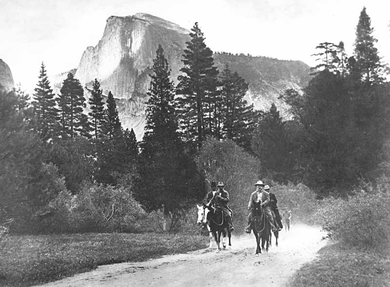 President Theodore Roosevelt and John Muir riding horses along a road in Yosemite Valley, with Half Dome in the distance, accompanied by Park Rangers Archie Leonard and Charles Leidig, followed by unidentified man on foot; left to right, Leonard, Muir, Roosevelt, Leidig